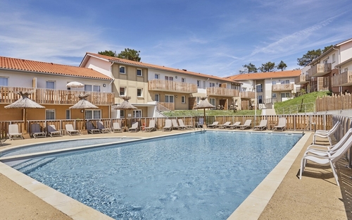 Poolbereich einer Ferienresidenz mit Liegestühlen und Sonnenschirmen vor Gebäuden.