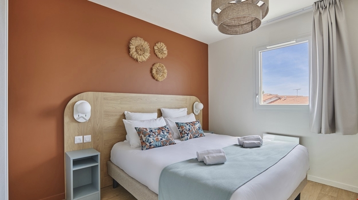 Schlafzimmer mit Doppelbett, oranger Wand, Fenster und dekorativen Wandblumen.