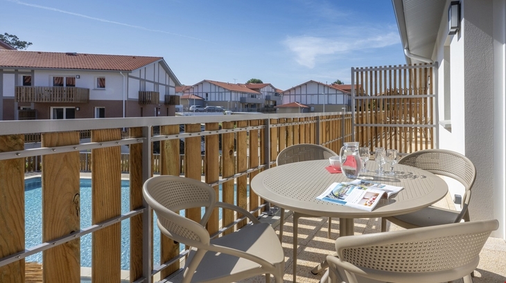 Balkon mit Tisch, Stühlen und Getränken, Blick auf Pool und Gebäude im Hintergrund.