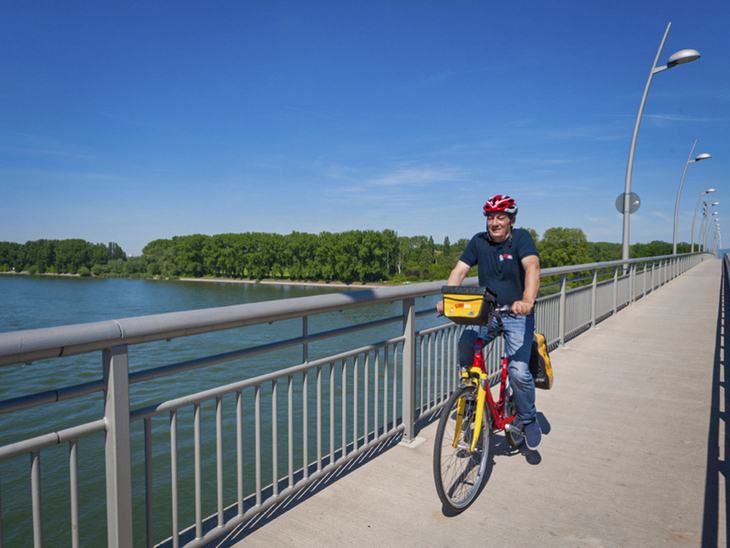 Radfahrer auf einer Brücke am Rhein bei sonnigem Wetter.