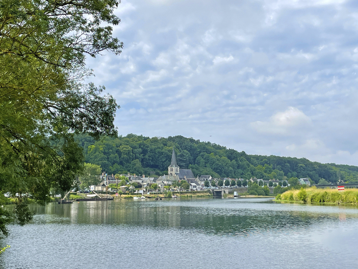 Flusslandschaft mit Dorf und Kirche im Hintergrund unter bewölktem Himmel.
