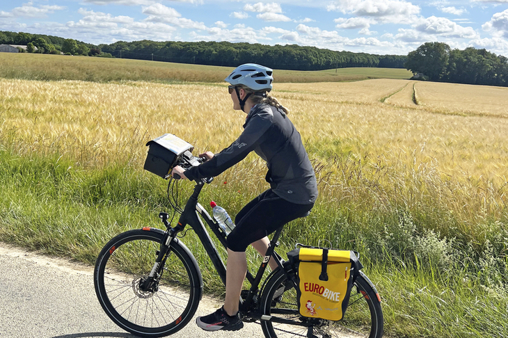 Radfahrer auf Landstraße neben Getreidefeld bei sonnigem Wetter.
