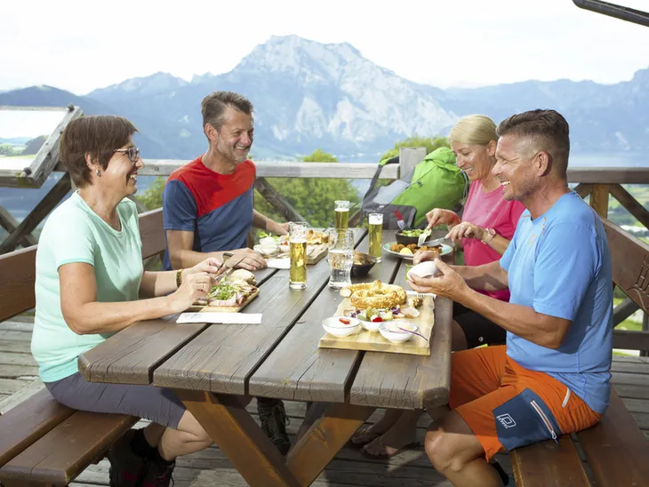 Personen essen gemeinsam an einem Holztisch mit Bergblick im Hintergrund.