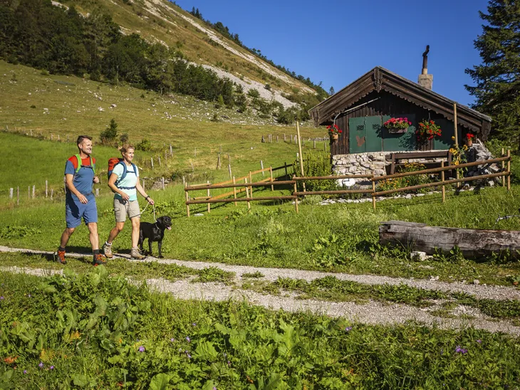 Zwei Menschen wandern mit einem Hund auf einem Pfad nahe einer Hütte im Salzkammergut.