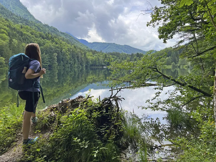 Wanderer steht am Ufer eines Sees im Salzkammergut, umgeben von Wald und Bergen.