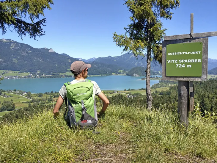 Person sitzt am Aussichtspunkt mit Blick auf den Wolfgangsee und Berge.