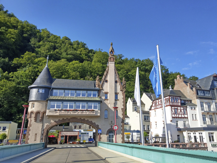 Stadttor im Jugendstil mit Fahnen und bewaldetem Hügel im Hintergrund