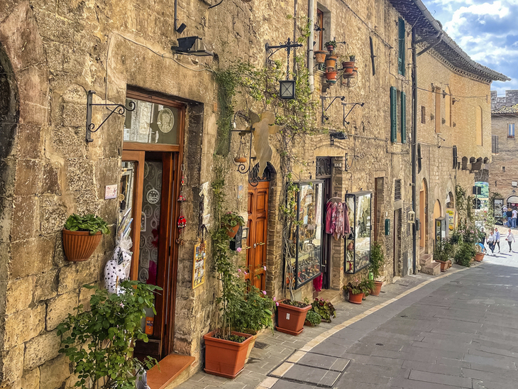 Schmale gepflasterte Gasse in Assisi mit historischen Steinhäusern und Blumendekorationen.