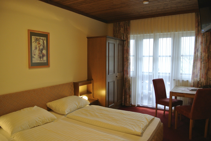 Hotelzimmer mit Doppelbett, Wandschrank, Tisch und Stühlen, Fenster mit Gardinen.
