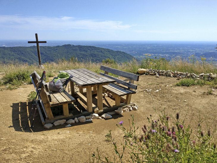 Holztisch mit Bänken und Kreuz vor Berglandschaft und Meer im Hintergrund.