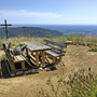 Holztisch mit Bänken und Kreuz vor Berglandschaft und Meer im Hintergrund.