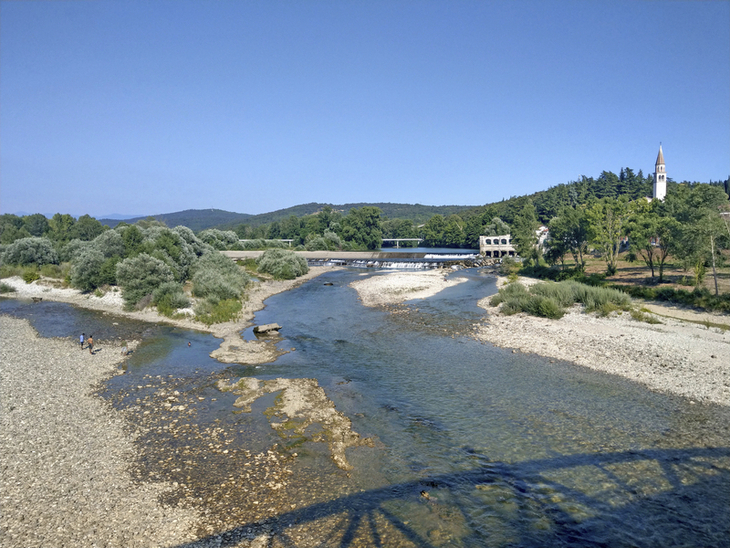 Flusslandschaft mit Brücke, Bäumen und Kirchturm im Hintergrund an einem sonnigen Tag.