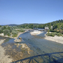 Flusslandschaft mit Brücke, Bäumen und Kirchturm im Hintergrund an einem sonnigen Tag.