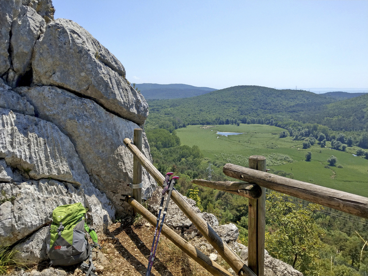 Wanderpfad mit Rucksack und Wanderstöcken, Blick auf grüne Hügel und Felsen.