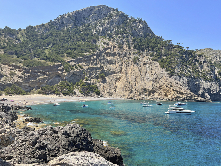 Boote im blauen Wasser vor bergiger Küste mit sandigem Strand und bewachsener Felswand.