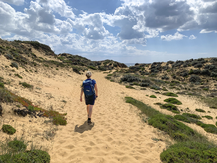 Person wandert durch sandige Dünenlandschaft unter blauem Himmel.