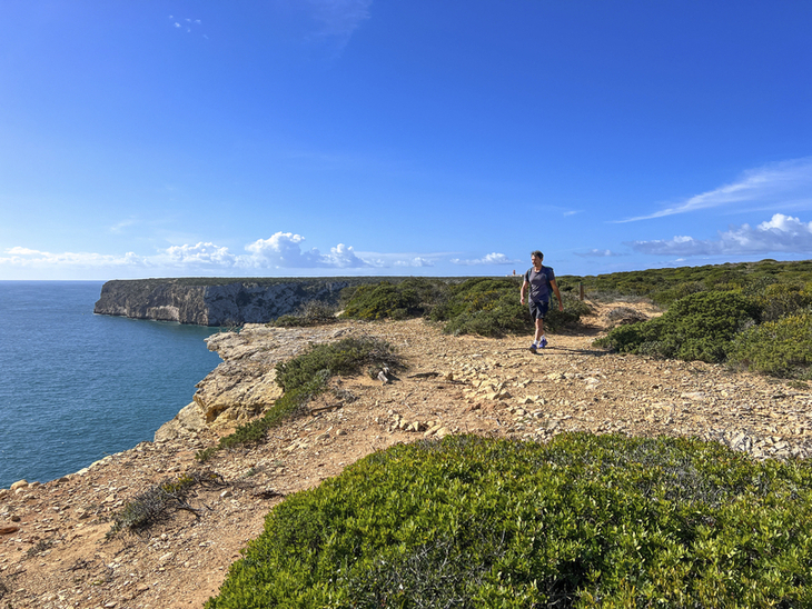 Person wandert an einer felsigen Küste mit Meerblick und blauem Himmel.