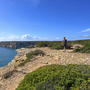 Person wandert an einer felsigen Küste mit Meerblick und blauem Himmel.