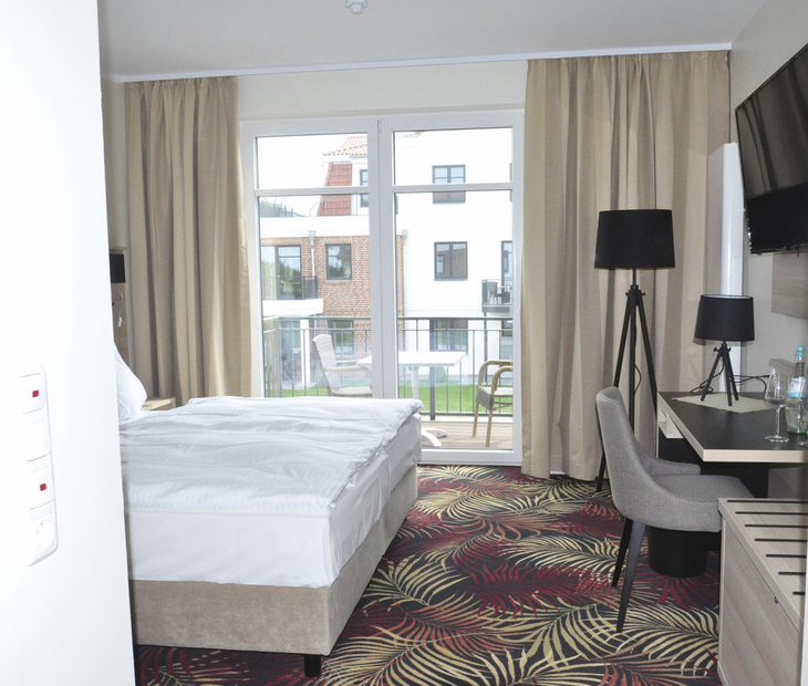 Hotelzimmer mit Bett, Schreibtisch, Balkon und Blick auf andere Gebäude.