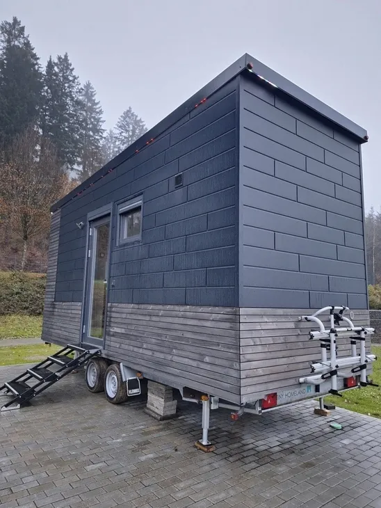 Kleines Haus auf Rädern mit dunkler Holzverkleidung, auf einem gepflasterten Platz.