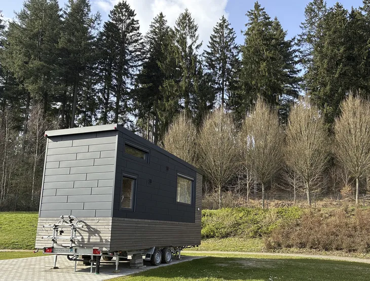 Kleines Tiny Haus auf Rädern vor einem Waldstück.