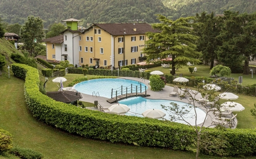 Garten mit Pool und Sonnenschirmen vor einem Hotel, umgeben von Bergen.