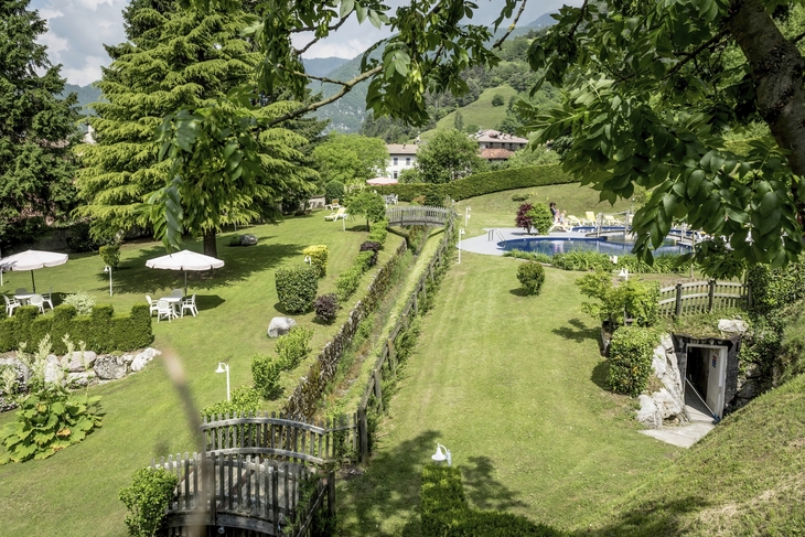 Hotelgarten mit Pool und Liegewiese, umgeben von Bäumen und Hügeln.