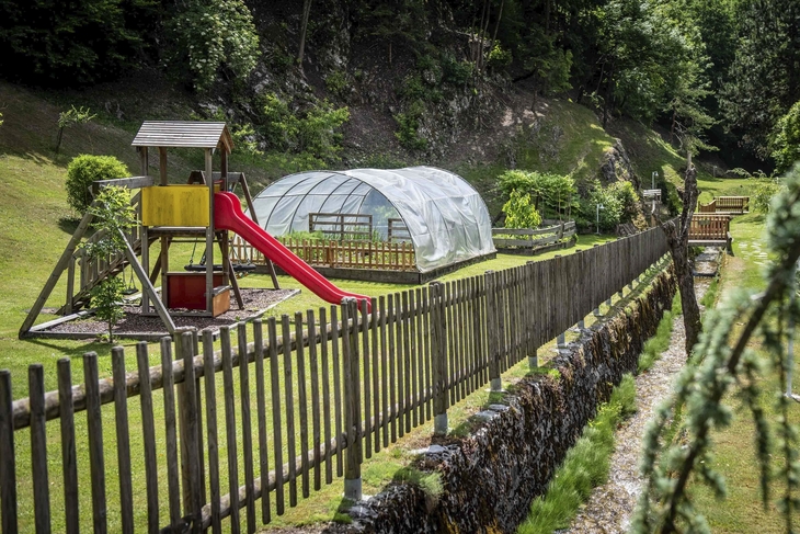 Spielplatz mit Rutsche und Gewächshaus in einem Garten, umgeben von Zaun und Bäumen.