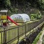 Spielplatz mit Rutsche und Gewächshaus in einem Garten, umgeben von Zaun und Bäumen.