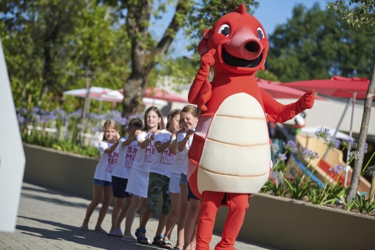 Kinder stehen lachend in einer Reihe hinter einem roten Maskottchen im Park Umag.