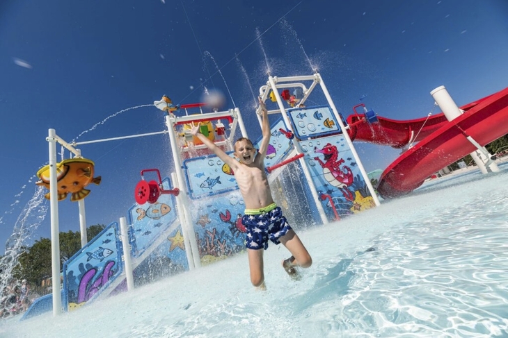 Kind springt in Pool bei Wasserspielplatz mit Rutsche und Sprinklern.