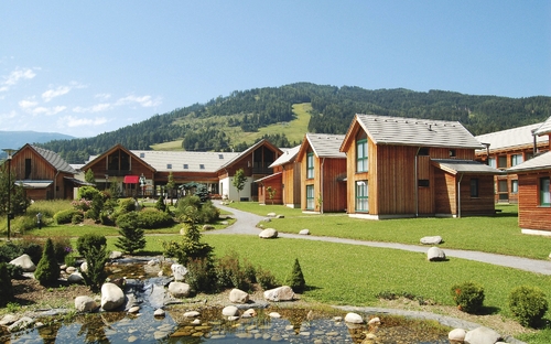 Ferienpark mit Holzhäusern und Teich vor bergiger Landschaft im Sommer.