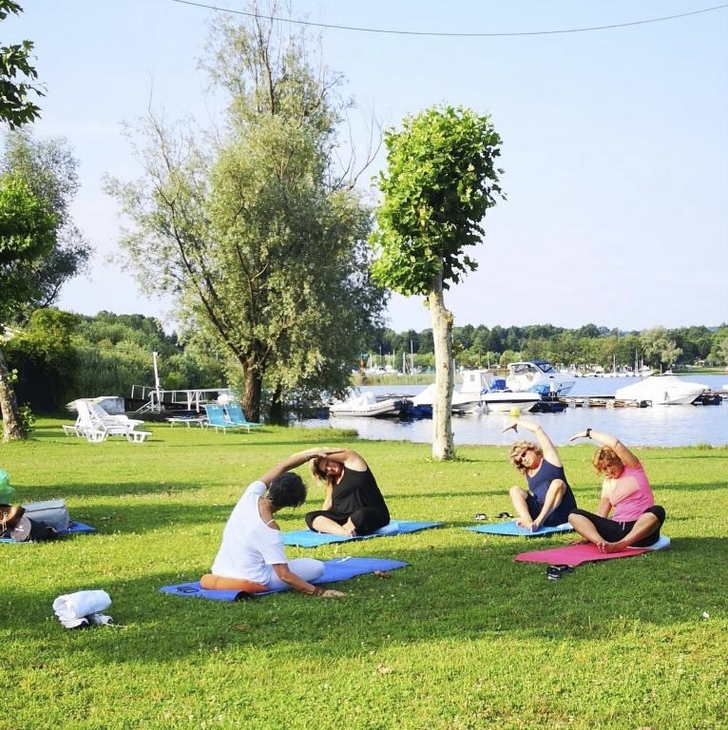Personen machen Yoga auf einer Wiese nahe einem Fluss oder See mit Booten im Hintergrund.
