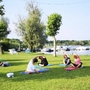 Personen machen Yoga auf einer Wiese nahe einem Fluss oder See mit Booten im Hintergrund.