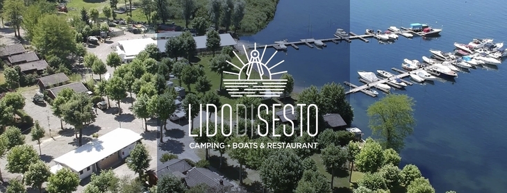 Luftaufnahme eines Campingplatzes mit Bäumen und angrenzendem See mit Bootsstegen.