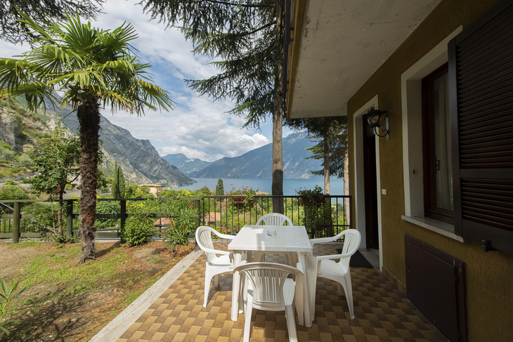 Terrasse mit Tisch und Stühlen, umgeben von Garten, Blick auf Berge und See.
