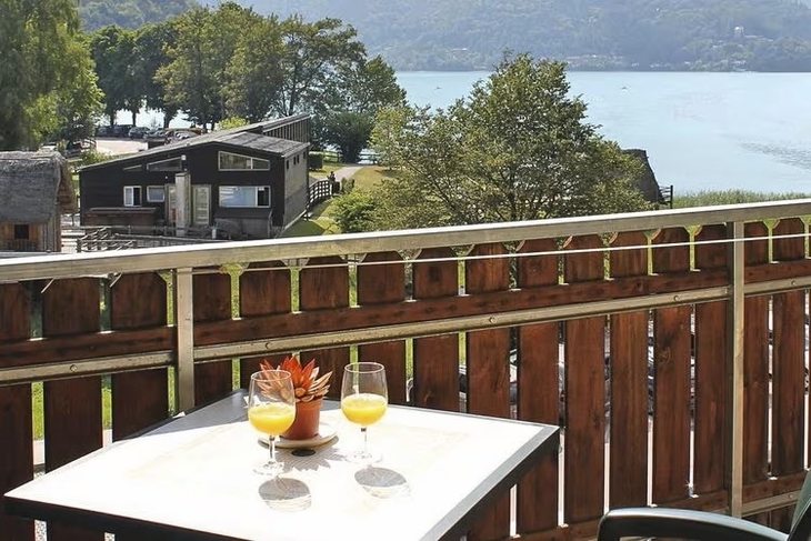 Blick von Balkon auf See und Hügel, zwei Gläser mit Getränken auf Tisch.
