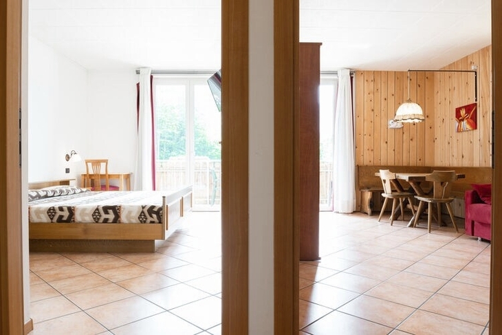 Zwei Räume mit Holzmöbeln und großen Fenstern; Schlafzimmer links, Wohnzimmer rechts.