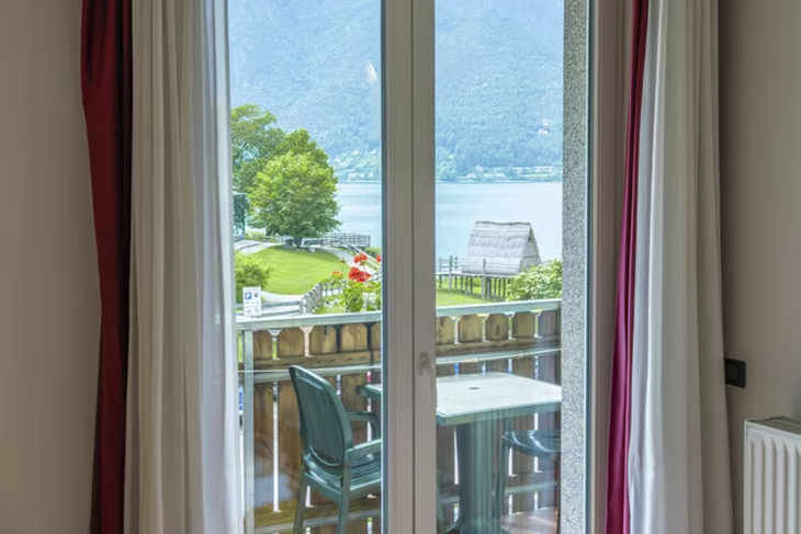 Blick durch ein Fenster auf einen Balkon mit See- und Berglandschaft im Hintergrund.