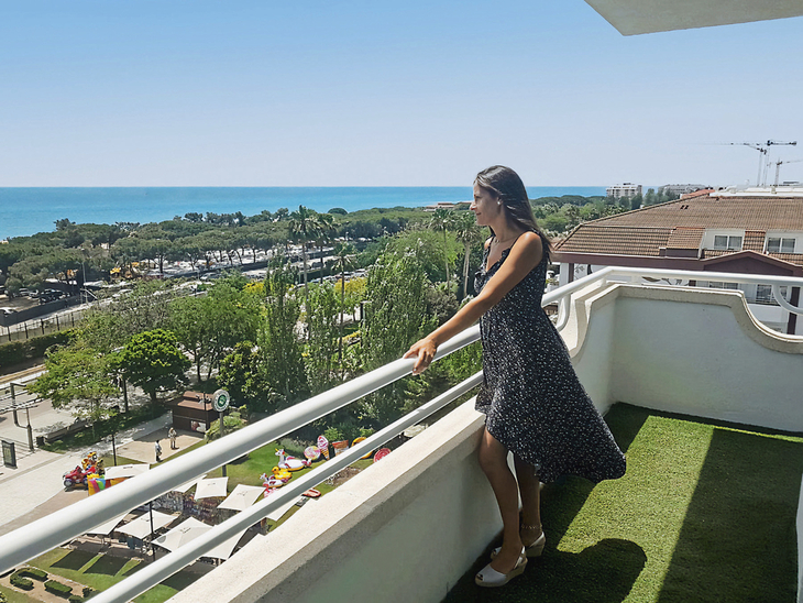 Frau auf Balkon mit Meerblick und grüner Landschaft im Hintergrund.