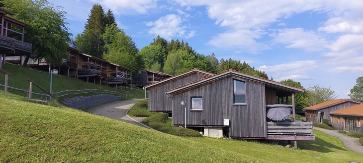 Holzhütten auf einem grünen Hügel, umgeben von Bäumen und blauem Himmel.