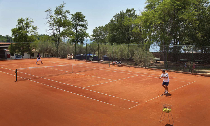 Tennisspiel auf einem Sandplatz mit zwei Spielern und Bäumen im Hintergrund.