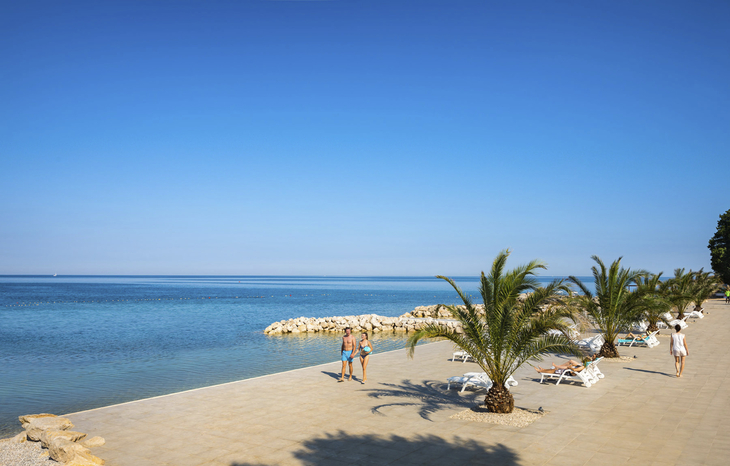 Promenade am Meer mit Palmen und Menschen, klarer Himmel, ruhige Küste.