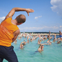 Person leitet Wassergymnastik in einem Pool mit vielen Teilnehmern unter blauem Himmel.