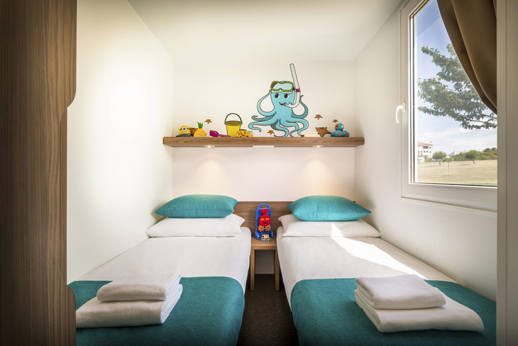 Zwei Einzelbetten mit blauen Decken, Spielsachen auf Regal, Fenster mit Blick nach draußen.