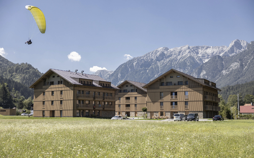 Drei Gebäude des Dormio Aparthotel Hinterstoder vor Bergkulisse im Sommer; Gleitschirmflieger.