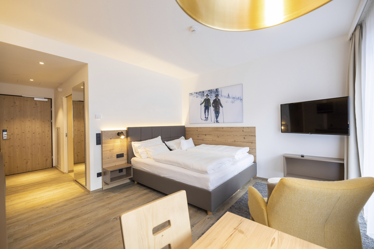 Helles Hotelzimmer mit Doppelbett, TV, Sessel und Holztisch.