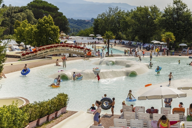 Menschen schwimmen und entspannen in einem Wasserpark mit Rutschen und Liegestühlen.