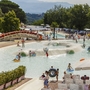 Menschen schwimmen und entspannen in einem Wasserpark mit Rutschen und Liegestühlen.