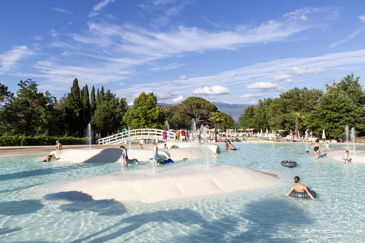Menschen genießen einen sonnigen Tag in einem großen, flachen Pool mit Wasserspielen.
